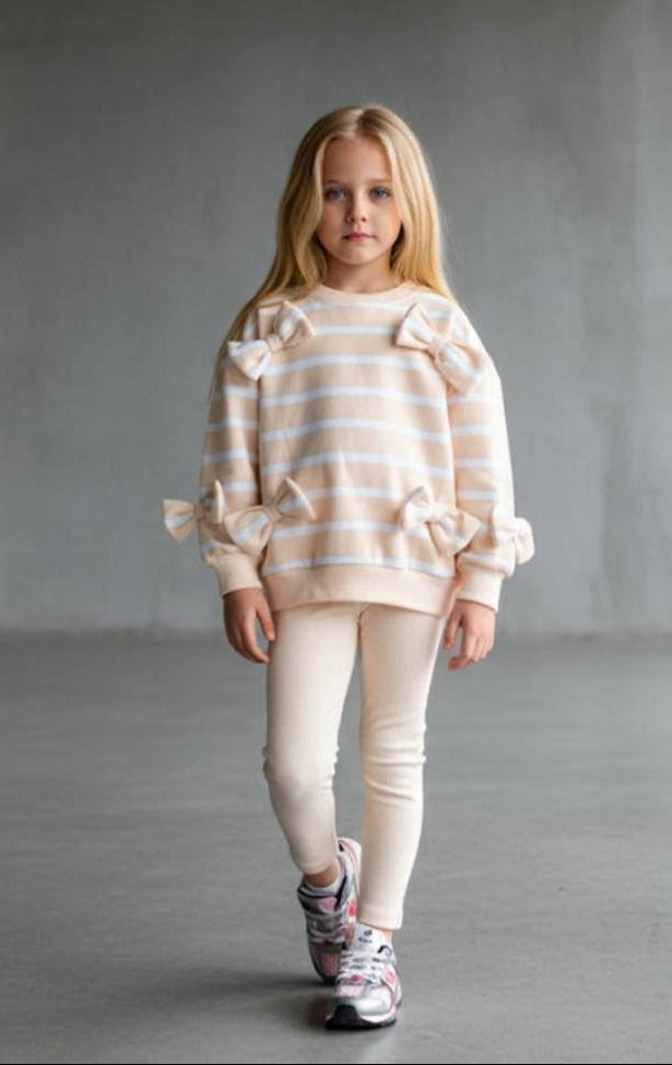 A young girl with blonde hair stands in a minimalist concrete room wearing a cream and light orange striped sweater with ruffle details on the shoulders and sleeves, paired with cream leggings and patterned sneakers.