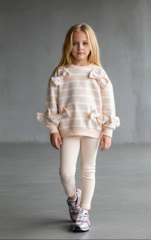 A young girl with blonde hair stands in a minimalist concrete room wearing a cream and light orange striped sweater with ruffle details on the shoulders and sleeves, paired with cream leggings and patterned sneakers.